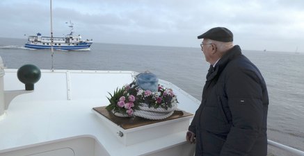 Älterer Mann steht auf einem Schiff, neben ihm eine blumengeschmückte Urne. Hintergrund: Meer und andere Schiffe