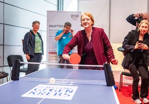 Im Vordergrund eine blaue Tischtennisplatte, mit der Aufschrift Bewegungen gegen Krebs. Die Seniorenministerin Lisa Paus spielt an der Platte Tischtennis. Sie trägt einen weinroten Blazer, darunter eine aubergine-farbene Bluse. Sie hat schulterlanges rötliches Haar. Um die Platte stehen Fotografen und andere Personen.