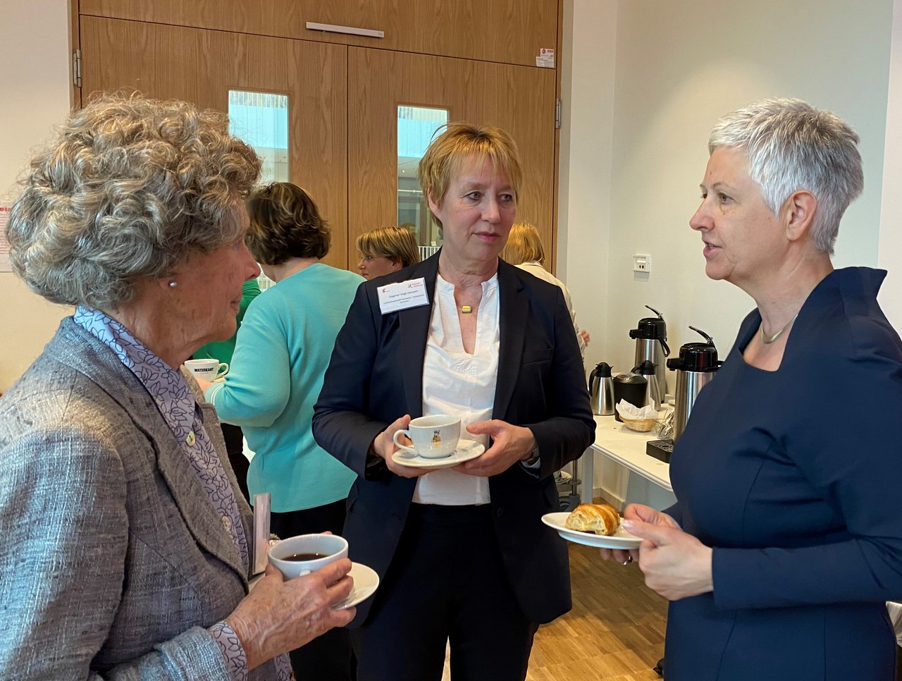 Drei Frauen mittleren Alters unterhalten sich im Stehen. Zwei Frauen halten eine Tasse Kaffee und eine Untertasse in der Hand. Eine Frau hält einen Teller mit einem Croissant in der Hand