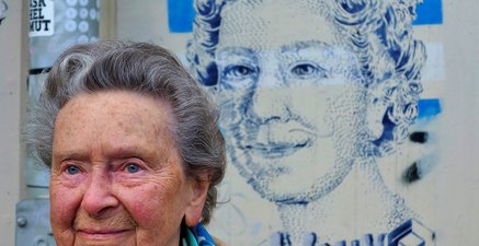 Zu sehen ist ein Graffiti der englischen Königin Elisabeth II. Davor steht eine Frau, die aussieht wie die Queen.