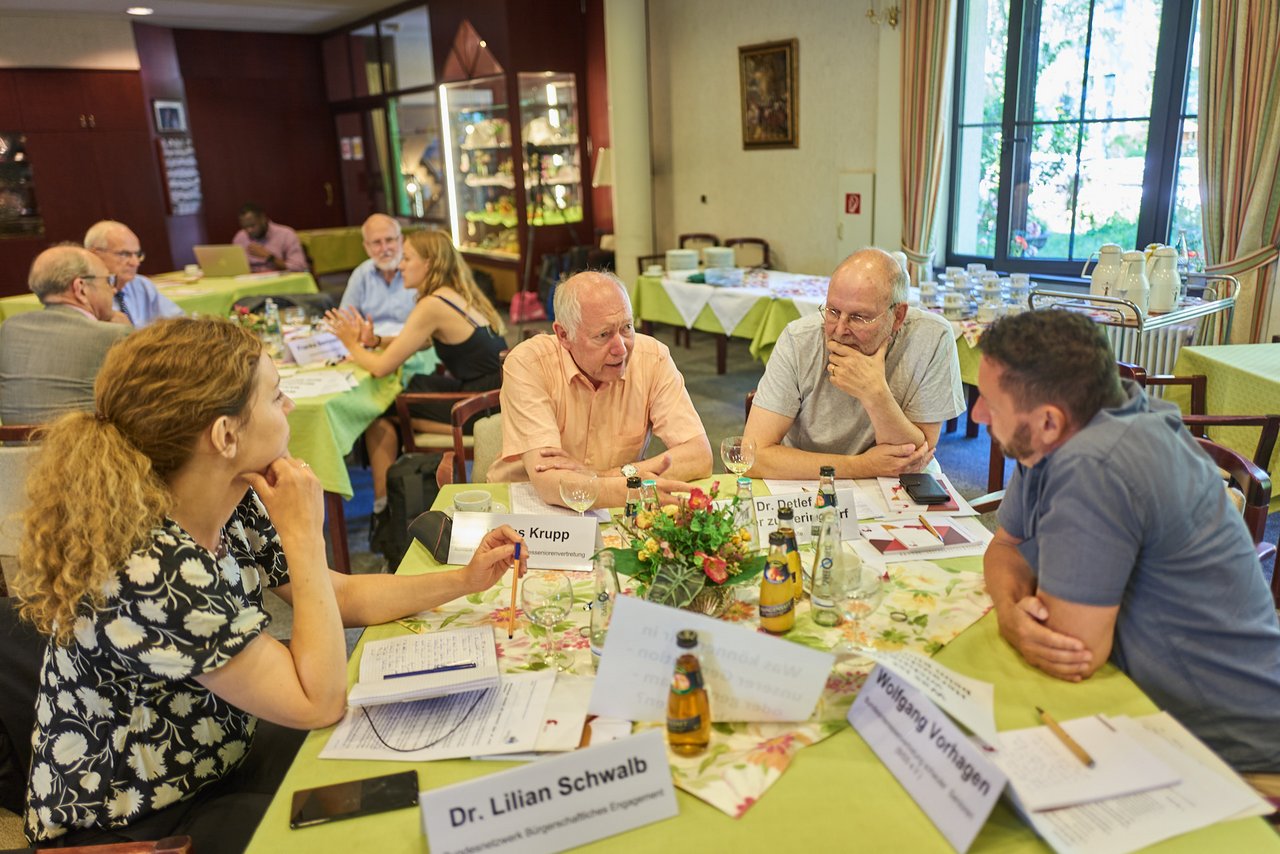 Im Veranstaltungsraum sind mehrere Tische mit neongrünen Tischdecken, an denen Menschen sitzen, die diskutieren. Am Tisch im Vordergrund sitzen zwei ältere, zwei Männer, und zwei jüngere Personen, ein Mann und eine Frau, die sich intensiv unterhalten.