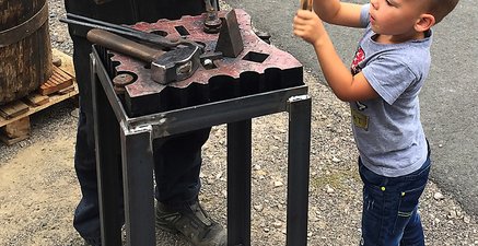 Arbeitsstand eines Schmiedes mit Werkstück und Werkzeug. Ein kleiner Junge schwingt einen Hammer, unter Aufsicht.  