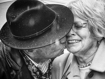 Schwarz-weiß Fotografie. Älterer Mann mit Hut küsst eine ältere Dame liebevoll im Regen.