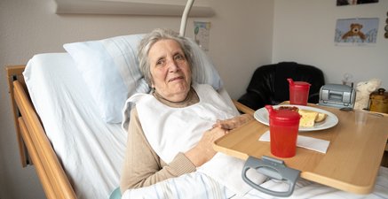 Alte, lächelnde Frau halb sitzend im Bett; kleiner Tisch vor ihr mit belegtem Brot und Trinkbecher, Bilder an Wand