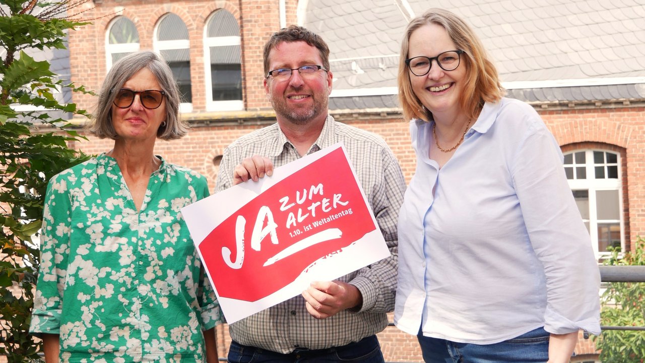 Zwei Frauen und ein Mann stehen im Freien. Der Mann hält ein Schild in der Hand mit der Aufschrift "Ja zum Alter!" Klick öffnet eine vergrößerte Ansicht.