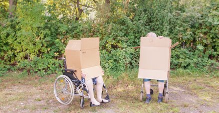 Senioren vor Bäumen, sie im Rollstuhl, er auf Rollator. Kartons bedecken Köpfe und Oberkörper.