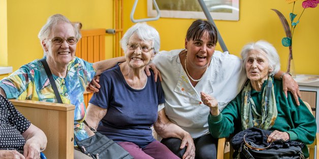 Ein Gruppe alter Menschen sitzen in einem gelb gestrichenen Raum auf einem Pflegebett zusammen. Neben dem Bett sitzt eine Frau im Rollstuhl und vor dem Bett eine Frau auf einem Stuhl. Zwischen ihnen sitzt eine Pflegerin in weißem Kittel. Sie haben die Arme auf den Schultern der Nachbarin gelegt und lachen.lten sich