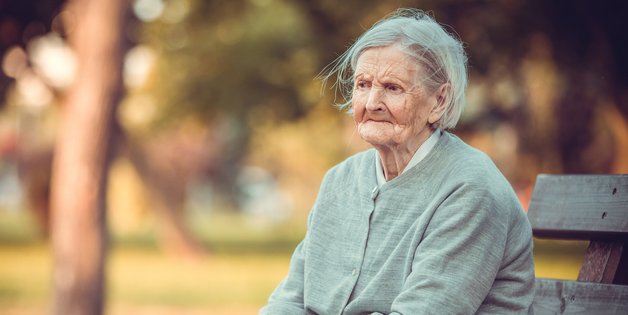 Eine alte Frau sitzt auf einer Bank im Park. Sie schaut sehr traurig