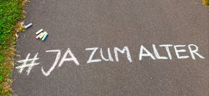 Mit weißer Kreide auf Asphalt geschrieben steht: "#JA zum Alter" Darüber liegen bunte Kreidestücke. 