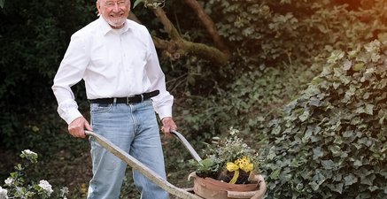 Schubkarre mit Blumenkübel darin wird von einem älteren Mann durch Garten geschoben