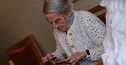 Eine weißhaarige, alte Frau hält Fotos in der Hand und schaut diese an. Sie trägt eine weiße Strickjacke.