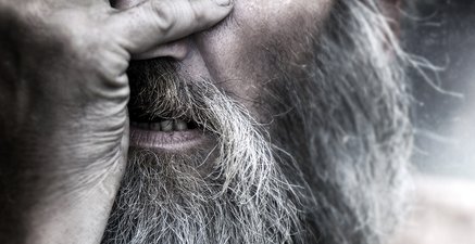 Älterer Mann mit Vollbart hält sich die Hand vor das Gesicht. Ein Auge schaut heraus.