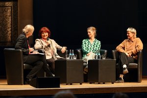 Bühne: In schwarzen Sesseln sitzen zwei junge Frauen, eine mittleren Alters und eine ältere Frau. Sie spricht gerade in ein Mikrofon.