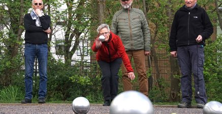 Frau wirft in gebückter Haltung eine Bowl-Kugel. 3 Männer beobachten mit unterschiedlicher Mimik