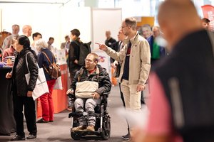 Ein Blick auf auf das Messegeschehen. Im Fokus, ein junger Mann im Rollstuhl. Neben ihm ein anderer junger Mann, der auf etwas zeigt.