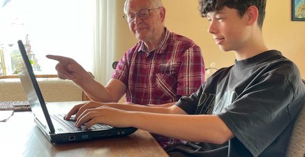 Zwei Männer sitzen am Tisch und schauen auf einen Laptop. Der ältere zeigt auf den Bildschirm, der jüngere tippt.