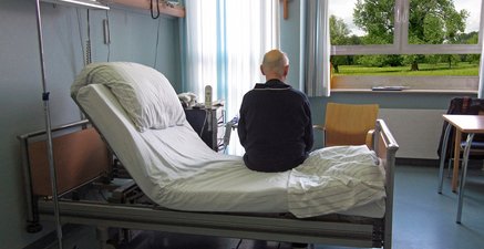 Älterer Mann sitzt im Schlafanzug auf einem Krankenhausbett und schaut zum Fenster, Rücken zur Kamera