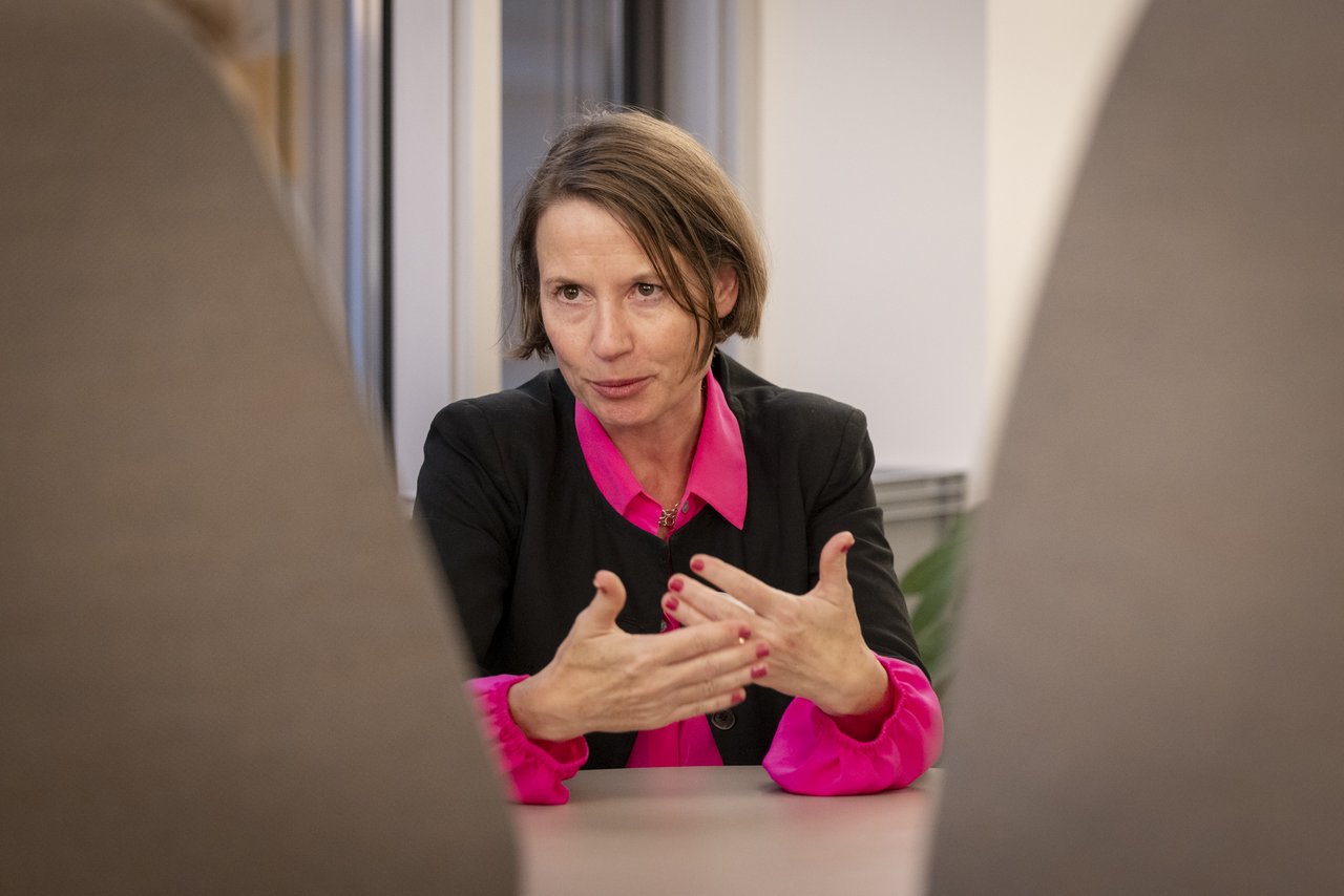 Eine Frau mittleren Alters sitzt an einem Tisch. Sie trägt einen schwarzen Blazer, darunter eine pinkfarbene Bluse. Sie spricht zu zwei Personen, die ihr gegenüber sitzen und gestikuliert mit beiden Händen. Klick öffnet eine vergrößerte Ansicht.