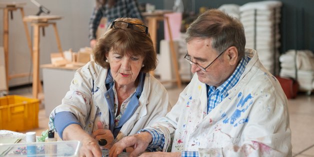 Ein älterer Mann und eine ältere Frau tragen Malerkittel, sitzen an einem Tisch und werken