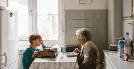 Hochaltrige Frau sitzt am Küchentisch und isst. Ihr Enkel gegenüber schreibt mit einer alten Schreibmaschine.