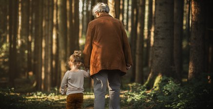 Von hinten: Ein kleines Mädchen spaziert an der Hand einer älteren Frau durch den Wald.