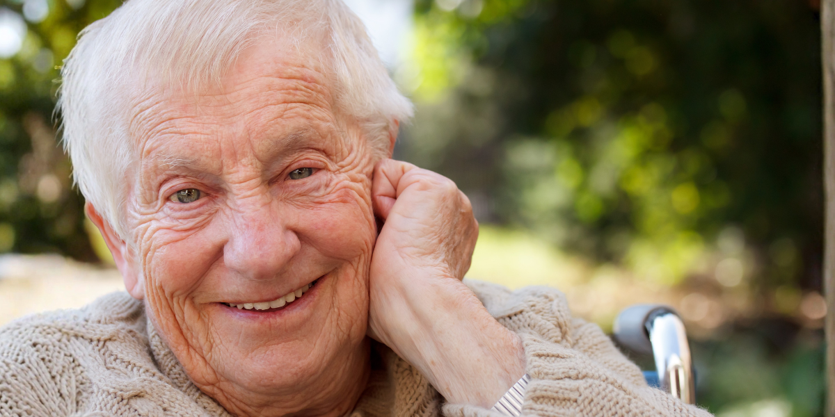 Alter Mann im Rollstuhl. Er sitzt draußen und lächelt in die Welt.