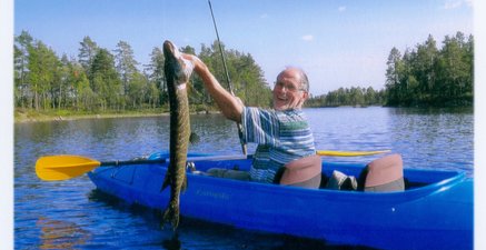 Älterer Mann sitzt in einem blauen Kajak auf einem See und hält strahlend einen Hecht in der linken Hand