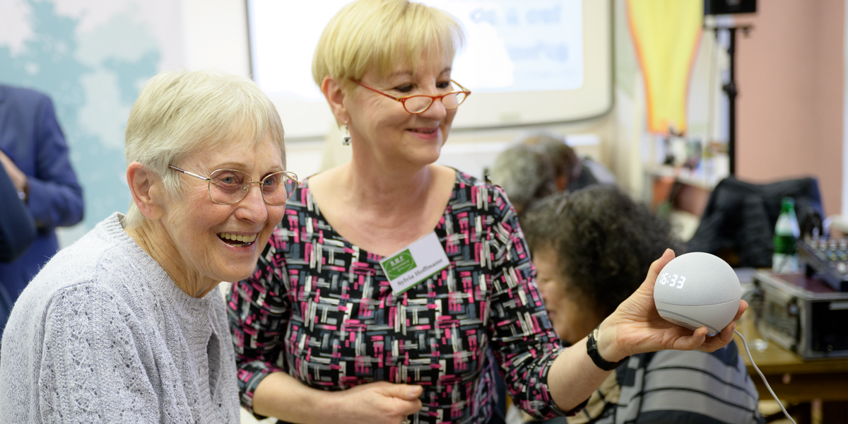 Drei ältere Frauen schauen sich gemeinsam ein digitales Gerät hat, das eine von ihnen in der Hand hält. Sie lachen.