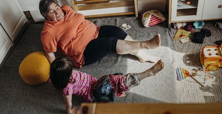 Eine ältere Frau und ein kleines Mädchen sitzen auf dem Fußboden eines Kinderzimmers und heben ein Bein in die Luft