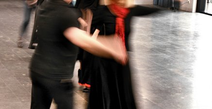 Ältere Frau tanzt mit einem älteren Mann Flamenco, CD-Spieler und Kursmitglieder im Hintergrund.