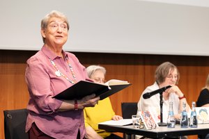 Eine ältere Frau mit rosa Bluse und einer bunten Kette. Sie hält eine schwarze Kladde in der Hand und ließt mit aufmerksamen Blick ins Publikum daraus vor.