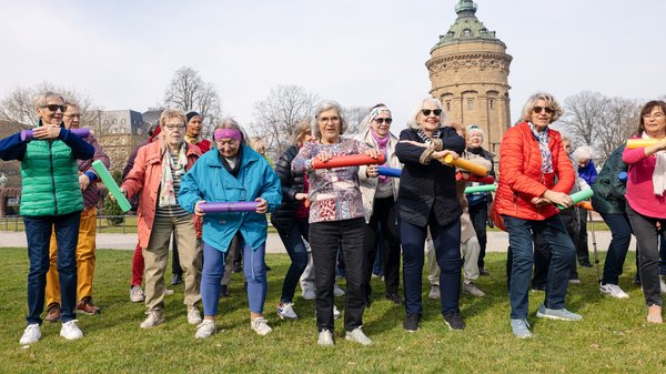 ©BAGSO_Lys Ältere Frauen stehen in einem Park. Sie haben in der Hand bunte Rollen und machen damit Gymnastik. Sie haben alle offensichtlich Spaß.