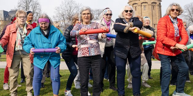 Ältere Frauen stehen in einem Park. Sie haben in der Hand bunte Rollen und machen damit Gymnastik. Sie haben alle offensichtlich Spaß.