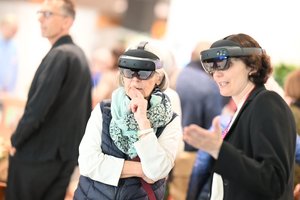 Eine junge und eine ältere Frau tragen außergewöhnliche Brillen. Sie sehen aus wir VR-Brillen. Sie schauen damit in die gleiche Brille.