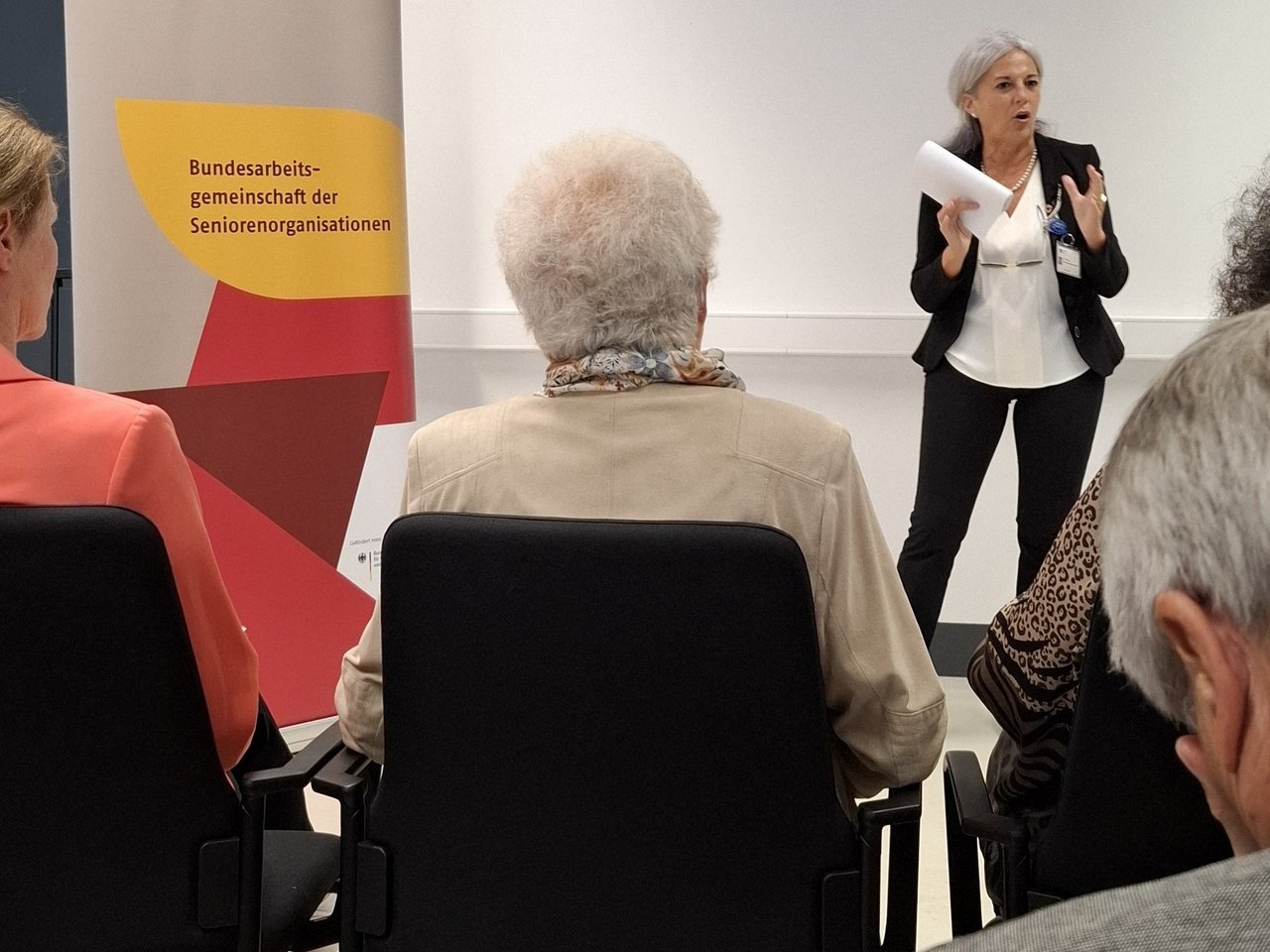 Ein Frau mittleren Alters spricht vor einer kleinen Gruppe. Neben ihr ist ein Roll Up mit der Aufschrift Bundesarbeitsgemeinschaft der Seniorenorganisationen zu sehen. Klick öffnet eine vergrößerte Ansicht.