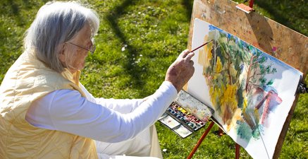 Das Bild zeigt eine alte Frau auf einer Wiese wie sie ein Landschaftsaquarell malt.