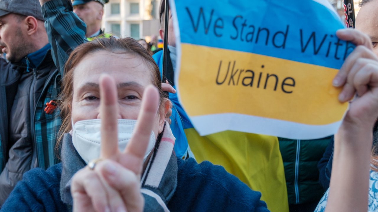 Eine ältere Frau hält ein Schild in der Hand. Auf dem ist die Flagge der Ukraine abgebildet, mit der Aufschrift "We stand with Ukraine". Mit der anderen Hand zeigt die Frau das Victory-Zeichen.