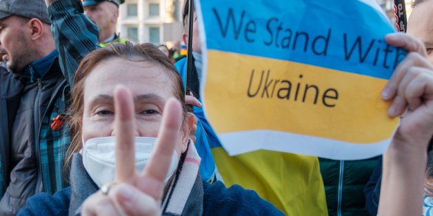 Eine ältere Frau hält ein Schild in der Hand. Auf dem ist die Flagge der Ukraine abgebildet, mit der Aufschrift "We stand with Ukraine". Mit der anderen Hand zeigt die Frau das Victory-Zeichen.