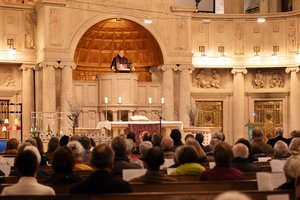 Ein Blick von hinten in einen Kirchraum. Ober auf der Kanzel steht ein Priester und spricht zu den Gläubigen.