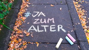 Auf dem Boden steht mit bunter Kreide geschrieben: "#JA zum Alter 1.1025 Weltaltentag." Unter dem Schriftzug liegen drei bunte Kreidestücke.