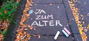 Auf dem Boden steht mit bunter Kreide geschrieben: "#JA zum Alter 1.1025 Weltaltentag." Unter dem Schriftzug liegen drei bunte Kreidestücke.
