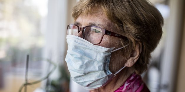 Ältere Frau mit Mund-Naschen-Schutz schaut aus dem Fenster