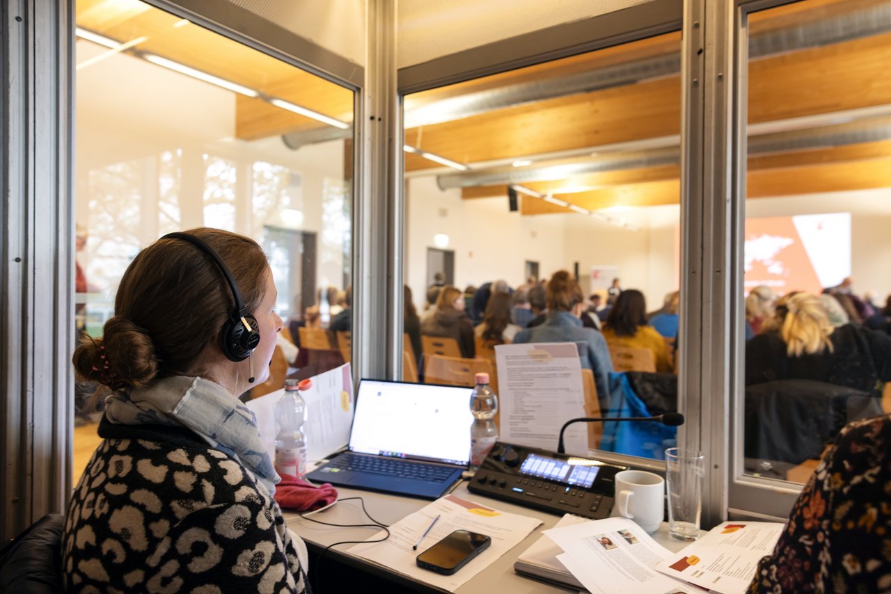 Eine Dolmetscherin mit Kopfhörern sitzt in einer Dolmetscherkabine. Vor ihr sind ein Laptop, die Verdolmetschungsanlage und verschiedene Unterlagen zu sehen. Durch die Glaswand der Kabine kann man in den Veranstaltungsraum mit dem Publikum sehen.