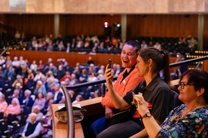 Ein voller Saal: Zwei Frauen sitzen auf einem Balkon. Eine der beiden hält ein Smartphone in der Hand und hält es Richtung Bühne. Sie lachen. Neben ihnen sitzt eine Frau mittleren Alters. Auch sie hält ihr Smartphone Richtung Bühne.