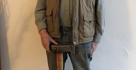Ein aufrecht stehender Mann in Arbeitskleidung, eine Axt steht mittig vor ihm, er lächelt erschöpft aber zufrieden.
