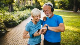 Ein älterer Mann und eine Frau stehen in einem Park. Sie haben beide Sportsachen an und tragen Kopfhörer. Die Frau hat ein Handy in der Hand und ihre Hand auf die Schulter des Mannes gelegt. Er trägt wahrscheinlich eine Smartwatch und zeigt auf das Zifferblatt.