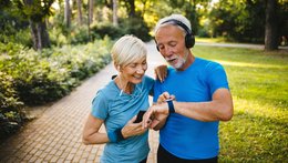 Ein älterer Mann und eine Frau stehen in einem Park. Sie haben beide Sportsachen an und tragen Kopfhörer. Die Frau hat ein Handy in der Hand und ihre Hand auf die Schulter des Mannes gelegt. Er trägt wahrscheinlich eine Smartwatch und zeigt auf das Zifferblatt.