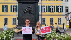 Ein Mann und eine Frau stehen vor einem Denkmal, von dem nur der Sockel zu sehen ist.  Sie halten beide jeweils ein Schild in der Hand mit der Aufschrift "Ja zum Alter!"