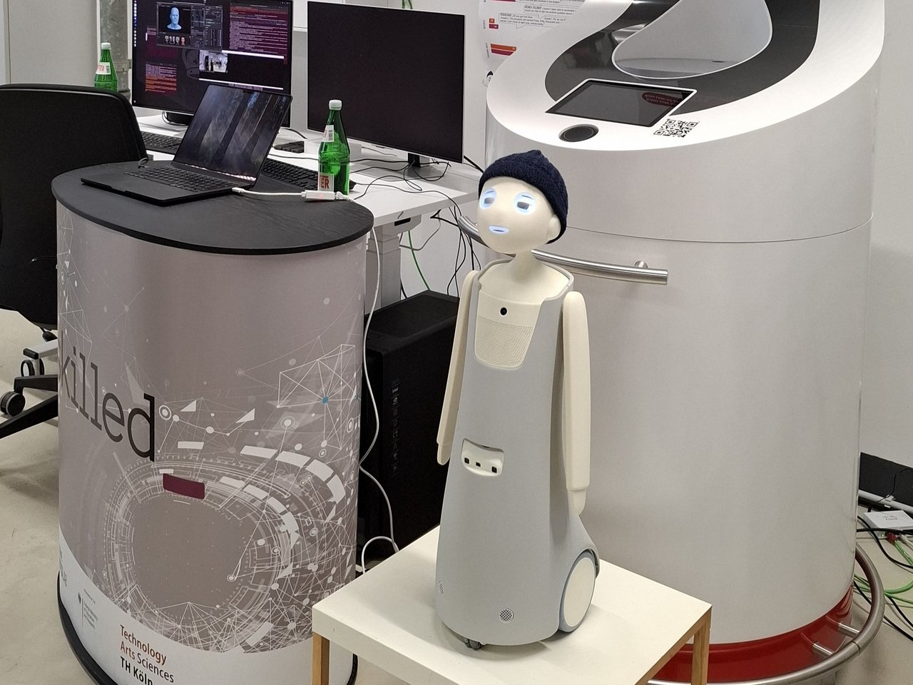 Ein kleiner Roboter mit Pudelmütze und Kulleraugen steht auf Rädern auf einem Tisch. Hinter ihm ist ein großes technisches Gerät zu sehen. Neben ihr ein Podest auf dem ein Laptop steht. Dahinter ist noch ein Schreibtisch, auf dem zwei Bildschirme zu sehen sind. Klick öffnet eine vergrößerte Ansicht.