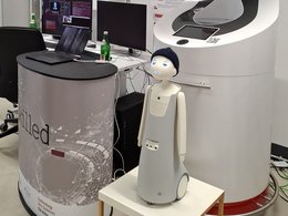Ein kleiner Roboter mit Pudelmütze und Kulleraugen steht auf Rädern auf einem Tisch. Hinter ihm ist ein großes technisches Gerät zu sehen. Neben ihr ein Podest auf dem ein Laptop steht. Dahinter ist noch ein Schreibtisch, auf dem zwei Bildschirme zu sehen sind.